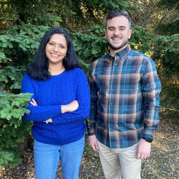 A man and a woman standing outdoors in front of green trees. The woman wears a bright blue cable-knit sweater and blue jeans, and the man wears a blue, brown, and gray plaid button-down shirt with beige pants.